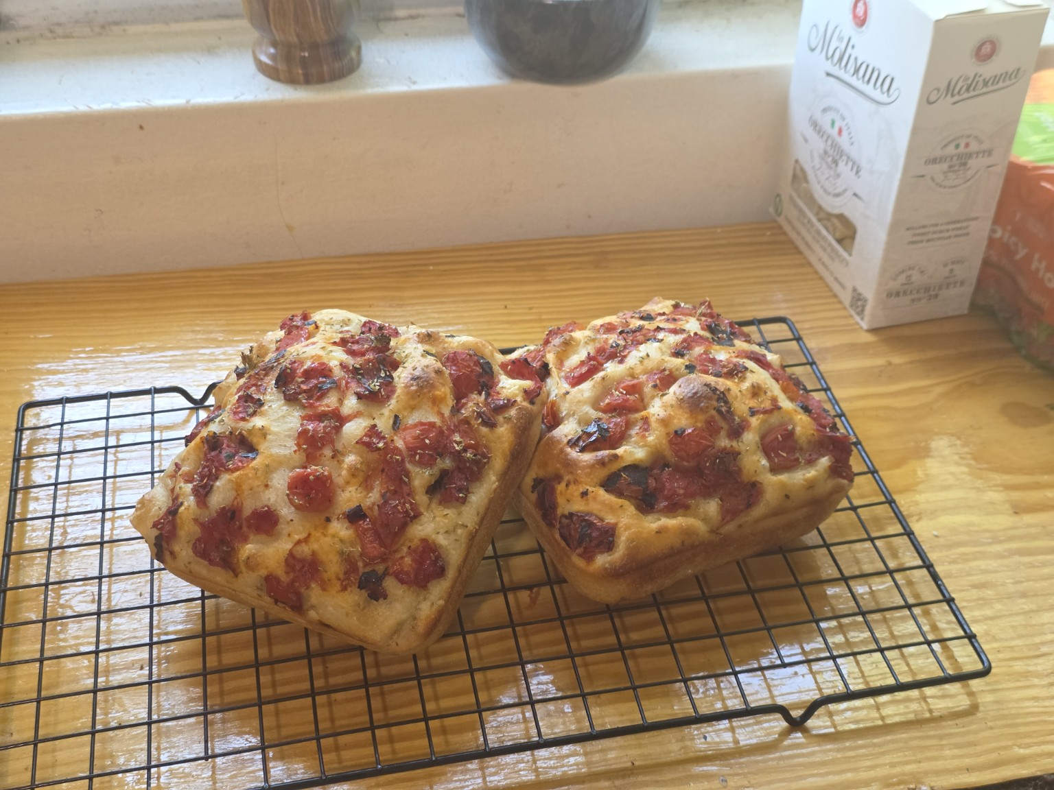 Two square tomato focaccias cooling on a wire rack. Both have risen into rounded, uneven domes well above the height of the pan, with roasted tomato pieces and oregano across golden tops.