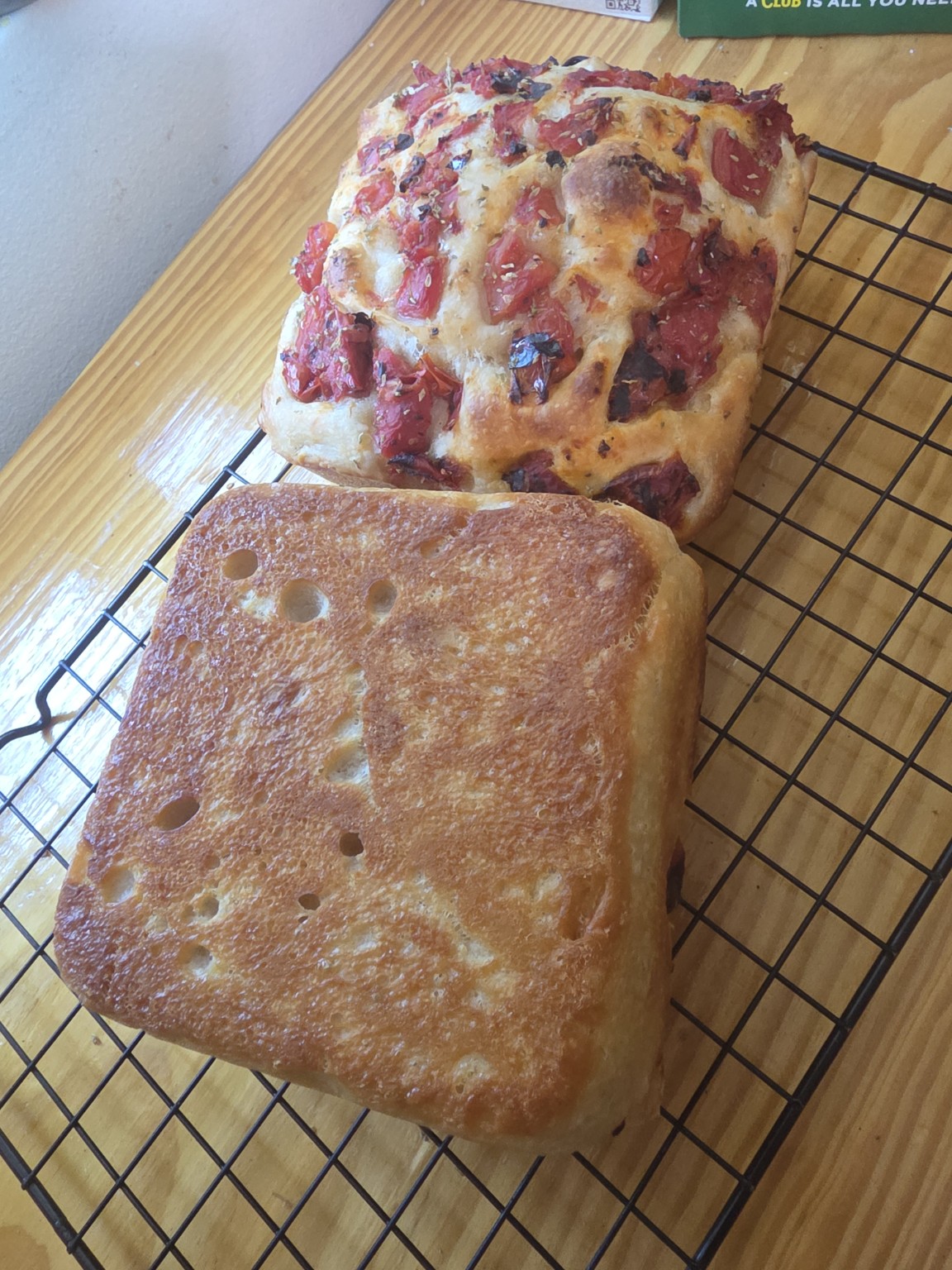 One loaf turned upside down on the rack, showing a deep, even golden-brown base with the dimple marks still visible. The second, tomato-topped loaf sits behind it.