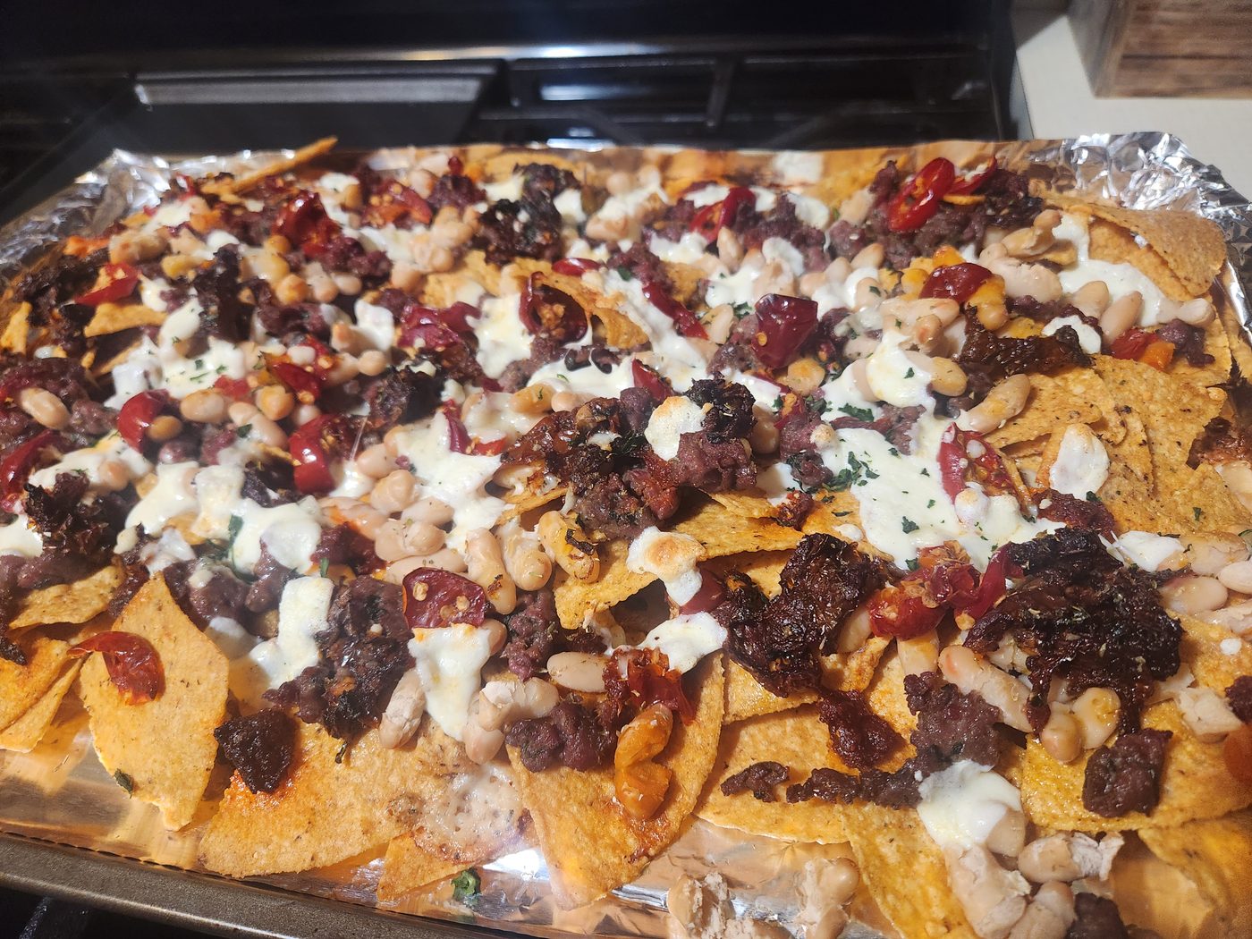 A foil-lined sheet pan of tortilla chips under melted mozzarella, whole cannellini beans, browned crumbled sausage, dark pieces of nduja and capuliato, and whole red Calabrian chillies.