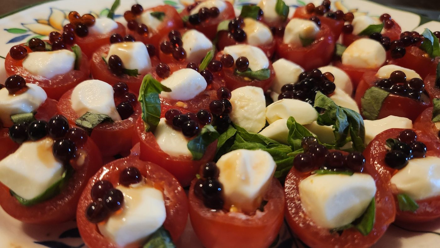 A close plate of halved cherry tomatoes, each hollowed and filled with torn basil and a piece of bocconcini, topped with clusters of glossy dark red vinaigrette pearls.