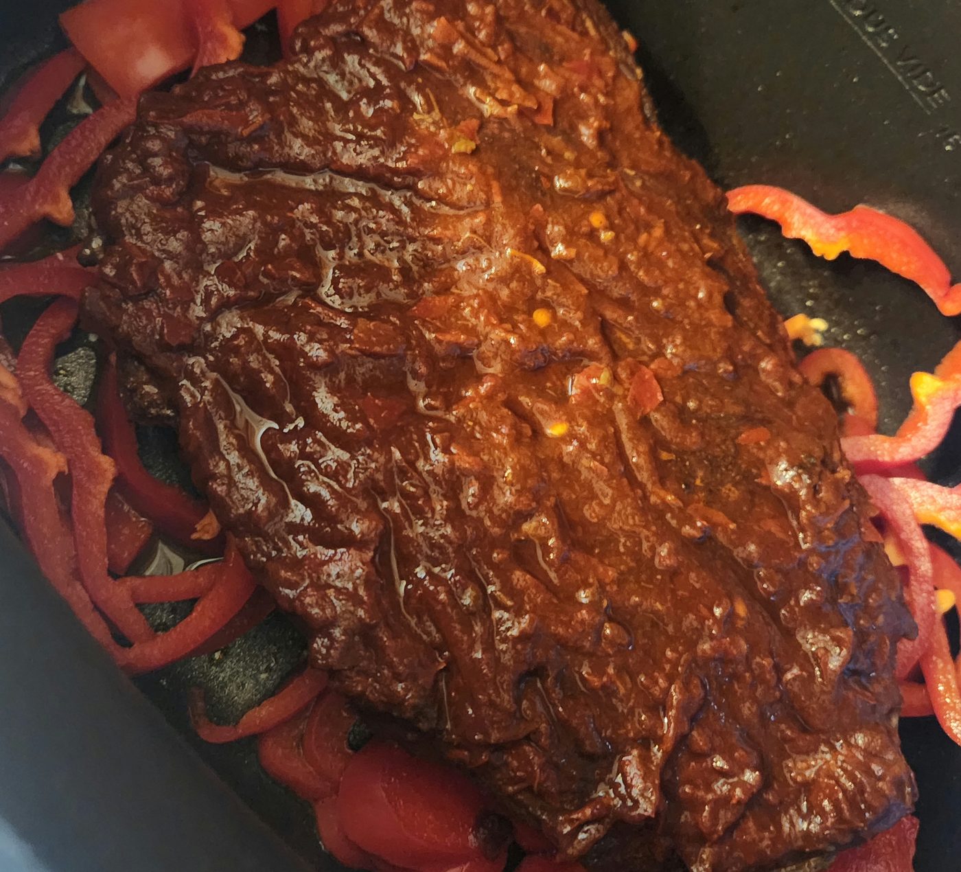 A rubbed roast in the crock pot on a bed of red pepper rings, coated in dark red chile slurry.