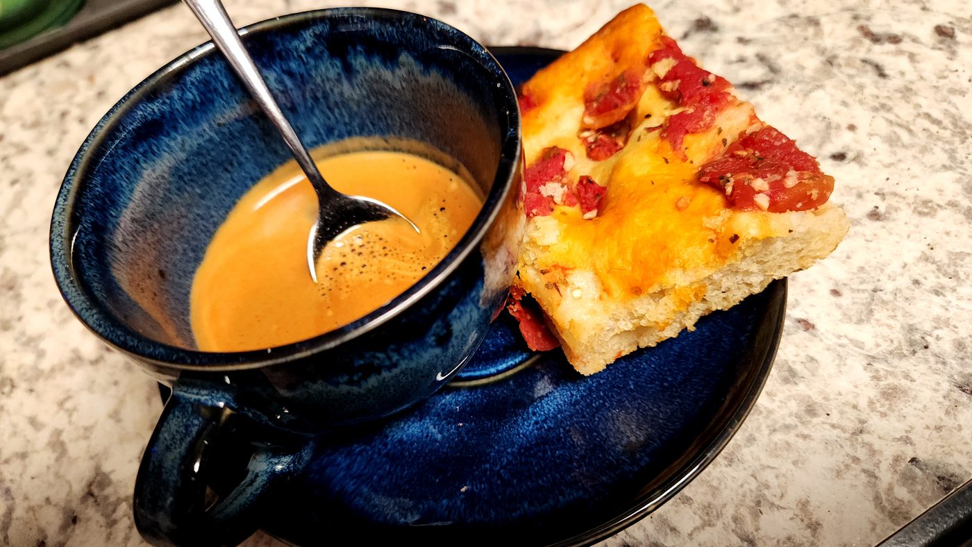 A double espresso in a dark blue cup with a slice of tomato focaccia on the saucer.