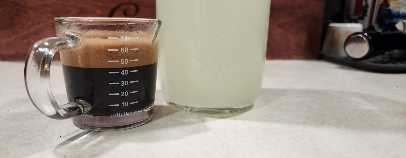 A jug of espresso beside a glass of lemonade on a white counter.