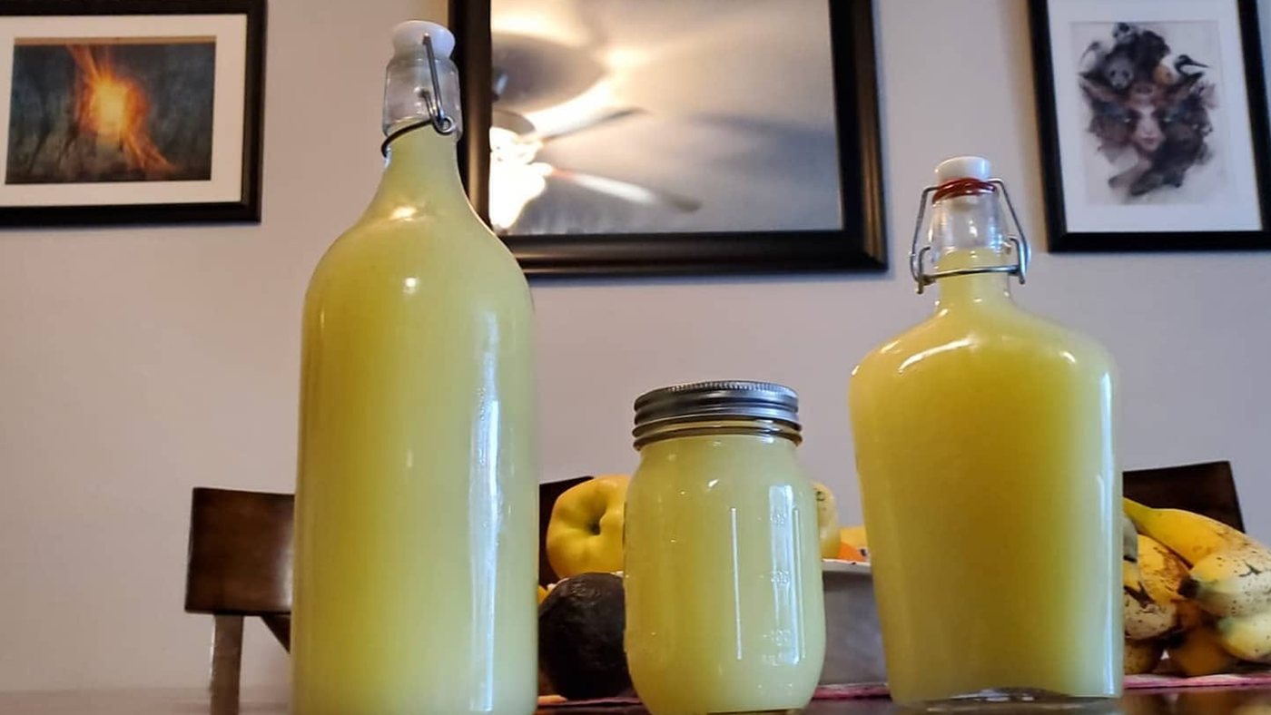 Two swing-top bottles and a mason jar of cloudy pale-yellow limoncello on a dining table, with a bowl of fruit behind them.